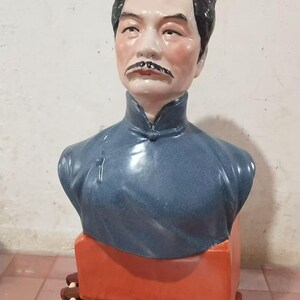 Może przedstawiać: Ceramiczne popiersie mężczyzny z ciemnymi wąsami i zaczesanymi do tyłu włosami. Ma na sobie niebieską tunikę. Popiersie stoi na czerwonej prostokątnej podstawie, wspartej na dekoracyjnej podstawie z drewnianych elementów.