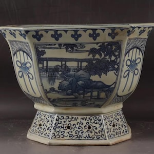 Op de afbeelding: Een blauw-witte keramische plantenbak met een achthoekige vorm. De plantenbak toont een landschap met bomen en een brug. Decoratieve patronen sieren de zijkanten en de basis, wat een elegante, traditionele uitstraling creëert.
