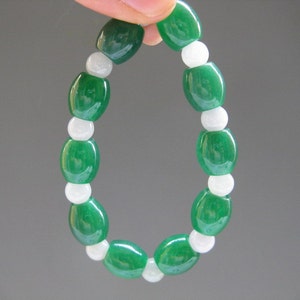 Puede incluir: Un brazalete de cuentas verdes y blancas. El brazalete presenta cuentas ovaladas verdes brillantes alternadas con cuentas redondas blancas. Las cuentas están ensartadas, formando un accesorio flexible y usable. El brazalete se muestra sobre un fondo neutro.