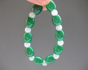 May include: A green and white beaded bracelet. The bracelet features oval-shaped, glossy green beads alternating with round, white beads. The beads are strung together, forming a flexible, wearable accessory. The bracelet is shown against a neutral background.