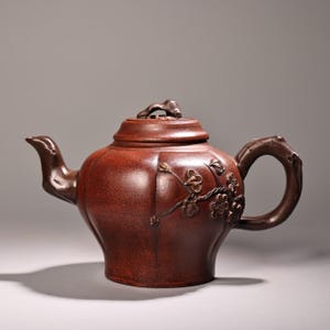 Puede incluir: Una tetera de arcilla de color marrón rojizo oscuro con una superficie texturizada y motivos en relieve de ramas de flor de ciruelo en el cuerpo. La tetera incluye un asa curva, un pico vertedor y una tapa con un pomo irregular.