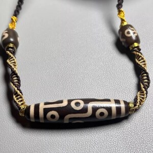以下が含まれることがあります： 白い幾何学模様が施された濃い茶色のビーズを中央に、小さなビーズを両側に配置したネックレス。ビーズは黒と金のツイストコードで繋がれており、ユニークでスタイリッシュなアクセサリーです。