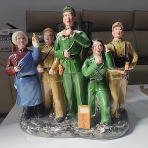 Może przedstawiać: Zestaw ceramicznych figurek przedstawiający pięć postaci w strojach wojskowych i tradycyjnych na skalistej podstawie. Centralna postać ma na sobie zielony mundur i trzyma lornetkę. Z przodu znajduje się mały pomarańczowy blok z chińskimi znakami.