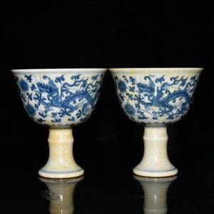Puede incluir: Dos copas antiguas de porcelana con diseños de dragones y flores azules. Las copas tienen una base y un tallo blancos con un cuenco estampado en azul y blanco. Las copas se exhiben sobre un fondo negro.
