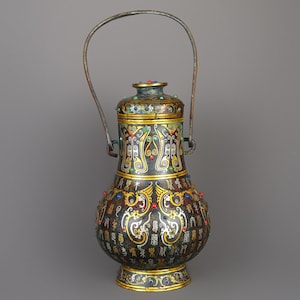 May include: An antique, ornate vase with a handle, featuring intricate gold and black designs. The vase is adorned with colourful gemstones and detailed calligraphy. The handle is made of a bronze-coloured metal. The vase has a bulbous shape and a decorative base.