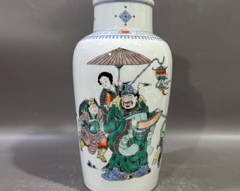 Op de afbeelding: Een witte keramische vaas met een brede body en een smalle hals. De vaas is versierd met een kleurrijke illustratie van figuren in traditionele kleding, waaronder een man met een rol en een ander onder een parasol. De rand heeft blauwe en rode accenten.