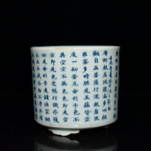 May include: A cylindrical white ceramic brush pot featuring blue Chinese calligraphy characters arranged in vertical columns around the exterior. The vessel has a smooth, glazed finish and rests on small, subtle feet at the base against a black background.