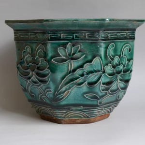 Peut inclure: Un pot de fleurs en céramique turquoise de forme hexagonale. Le pot présente des motifs floraux en relief et une bordure décorative. La base est de couleur brun-rouge, et la finition est brillante.