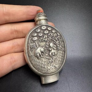May include: Antique silver-colored perfume bottle with a detailed relief design. The bottle features a scene with two figures, trees, and flowers. The stopper is topped with a small red stone. The bottle is oval-shaped and has a small base.