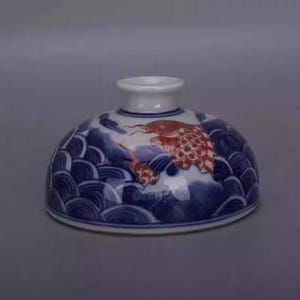 Könnte beinhalten: Eine Keramikvase mit rundem Boden und schmalem, zylindrischem Hals. Die Vase hat ein blau-weißes Wellenmuster und ein rot-weißes Fischdesign. Die Vase wird wahrscheinlich zu Dekorationszwecken verwendet.