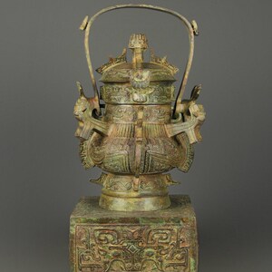 May include: Bronze vessel with intricate carvings and a handle. The vessel features detailed relief patterns and is mounted on a square base with ornate designs. The bronze has a weathered, aged appearance, with hints of green and brown.