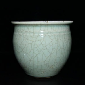 May include: A celadon ceramic pot with a rounded form and a gently outward-curving rim. The surface is marked with a network of fine cracks, creating a textured effect. The pot has a pale green colour and is set against a dark backdrop.