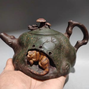 May include: A handcrafted, dark green teapot with a brown handle and spout. The teapot features a carved tiger emerging from a cave-like opening. The lid is adorned with mushroom and leaf details.