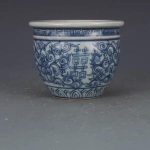 Könnte beinhalten: Ein kleiner, runder Keramiktopf mit weißem Rand und blau-weißem Blumendesign. Der Topf hat ein sich wiederholendes geometrisches Muster in der Nähe des Randes und ein zentrales Doppel-Glücks-Symbol. Der Boden ist hellbraun.