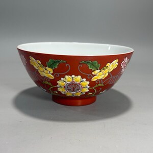 Puede incluir: Un cuenco de cerámica con un exterior de color rojo intenso que presenta un patrón pintado a mano de flores amarillas y blancas con hojas verdes. El centro del diseño floral incluye un emblema circular con un carácter rojo. El interior es blanco.