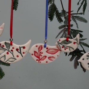 May include: A collection of hand-painted ceramic bird ornaments. The birds feature red, green, and white floral patterns and are suspended by ribbons. The ribbons are in shades of red, blue, and grey. The ornaments hang from a green branch.