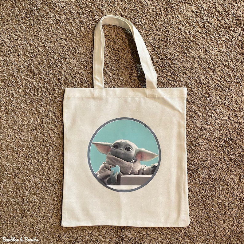 K&ouml;nnte beinhalten: Eine wei&szlig;e Canvas-Tragetasche mit einem runden Design, das eine Figur aus der Star Wars-Serie The Mandalorian zeigt. Die Figur ist in einem hellblauen Kreis mit grauer Umrandung dargestellt und h&auml;lt ein blaues Objekt in der Hand.