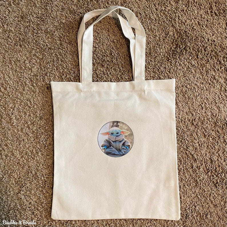 May include: A white canvas tote bag with a graphic of a character from the Star Wars franchise. The graphic is a circle with the text "It's a mood" and a picture of the character.