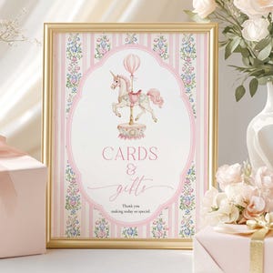 May include: A framed sign with a gold border, featuring a pink and white striped background and a carousel unicorn illustration. The sign reads "CARDS & gifts" and "Thank you making today so special."