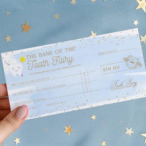 May include: A light blue check from "The Bank of the Tooth Fairy" with a cartoon tooth illustration. The check is for $10.00 and includes fields for the recipient's name and the amount in words. The check is held against a blue background with gold star decorations.