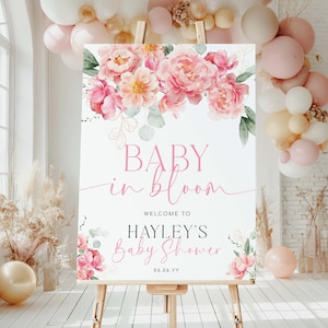 May include: A white welcome sign with pink floral illustrations and gold accents. The text reads "Baby in Bloom" and "Welcome to Hayley's Baby Shower 06.06.YY".
