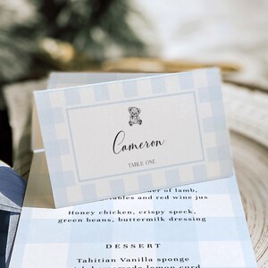 May include: A light blue and white checked place card with the name "Cameron" in cursive, a small teddy bear illustration, and the text "TABLE ONE". The card is part of a baby shower or event.