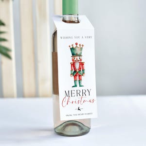 May include: A wine bottle with a green top and a white tag. The tag features a watercolor illustration of a nutcracker soldier with a crown, and the words "Wishing You a Very Merry Christmas" in red and black script. The tag also includes a reindeer graphic and the family name.