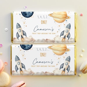 May include: Two chocolate bar wrappers with a space theme. Each wrapper features a rocket ship, planets, and stars. Text includes "5, 4, 3, 2... ONE!" and "Cameron's FRIST TRIP AROUND THE SUN!" in a whimsical font.