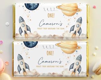 May include: Two chocolate bar wrappers with a space theme. Each wrapper features a rocket ship, planets, and stars. Text includes "5, 4, 3, 2... ONE!" and "Cameron's FRIST TRIP AROUND THE SUN!" in a whimsical font.