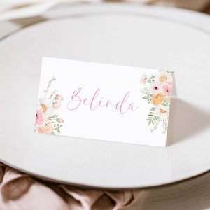 May include: A white place card with the name "Belinda" in pink script. Floral watercolor designs in shades of pink, peach, and white adorn the corners. The card rests on a white plate, suggesting a wedding or event setting.