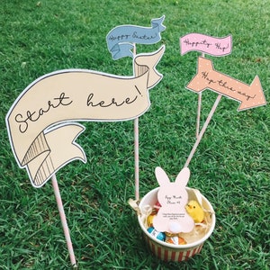 Può includere: Un gioco di caccia alle uova di Pasqua allestito sull'erba verde. Il gioco include un cartello "start here", un cartello "hop this way", un cartello "Happy Easter", un cartello "Happity Hop" e un cestino con un biglietto di indizio per la caccia alle uova e uova di cioccolato.