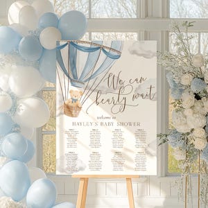 May include: A baby shower welcome sign with a watercolor hot air balloon design in shades of blue and white. The sign reads "We can bearly wait" and welcomes guests to Hayley's baby shower. A teddy bear is in the hot air balloon basket.