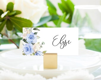 May include: A white place card with a watercolour design of blue flowers and greenery. The name "Elyse" is written in black cursive on the card. The card is held in place by a gold metal stand.
