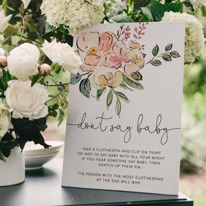 May include: A white card with a floral illustration and the text "don't say baby". The card has instructions for a game where guests try not to say the word "baby" and collect clothespins from those who do.