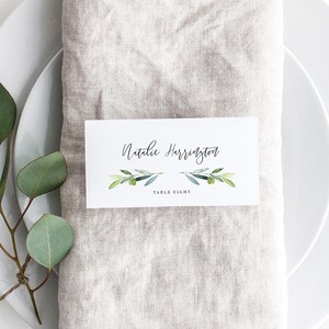 Può includere: Un segnaposto bianco con il nome "Natalie Harrington" in elegante corsivo, decorato con rami di ulivo ad acquerello. La carta mostra anche "TABLE EIGHT" in un semplice carattere, su un tovagliolo di lino e un piatto bianco.