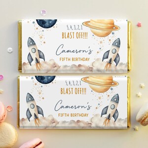 May include: A white chocolate bar wrapper with a gold border. The wrapper features a watercolour illustration of a rocket ship, planets, and stars. The text on the wrapper reads "5, 4, 3, 2, 1 BLAST OFF!!! Cameron's FIFTH BIRTHDAY".