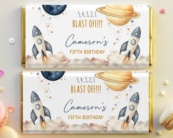 May include: A white chocolate bar wrapper with a gold border. The wrapper features a watercolor illustration of a rocket ship, planets, and stars. The text on the wrapper reads "5, 4, 3, 2, 1 BLAST OFF!!! Cameron's FIFTH BIRTHDAY".