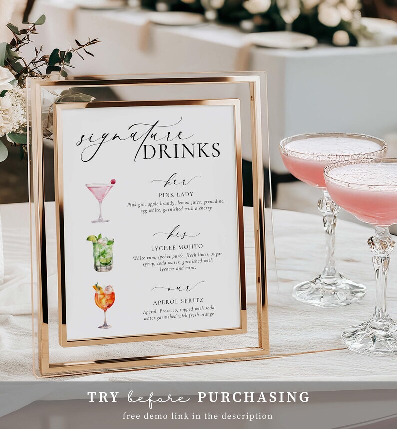 May include: A gold-framed sign with the words "Signature Drinks" and drink descriptions, including a Pink Lady, Lychee Mojito, and Aperol Spritz. Two pink cocktails in elegant glasses are in the background. The sign includes the text "TRY before PURCHASING".