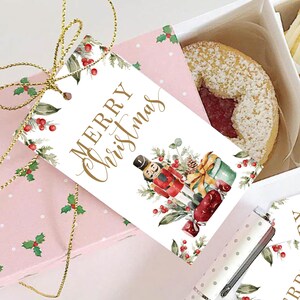 May include: A white gift tag with the text "Merry Christmas" in gold script. The tag features a watercolor illustration of a nutcracker, a pine cone, a wrapped gift, and red berries.