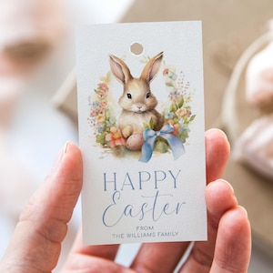 May include: A white gift tag with a watercolor illustration of a brown bunny rabbit holding a blue Easter egg. The bunny is surrounded by a wreath of colorful flowers. The text "Happy Easter" is written in blue script font. Below the text is the phrase "From The Williams Family".