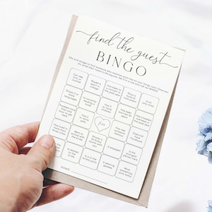 以下が含まれることがあります： 手で持たれた、黒い文字のベージュ色の「Find the Guest Bingo」カード。カードには、「2つ以上の言語を知っている」や「婚約している」などのプロンプトが書かれた正方形のグリッドがあります。このカードは、ブライダルシャワーや結婚披露宴のゲーム用に設計されています。