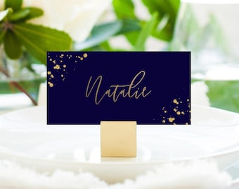 May include: A navy blue place card with gold glitter accents and the name "Natalie" written in gold calligraphy. The card is sitting on a gold metal stand.