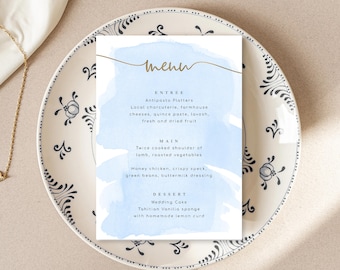 Puede incluir: Una tarjeta de menú de acuarela azul y blanca con letras doradas. El menú incluye entradas, platos principales y postres. El texto dice: "Menú", "Entrada", "Platos de antipasto", "Charcutería local, quesos de granja, pasta de membrillo, lavosh, fruta fresca y seca", "Plato principal", "Paletilla de cordero cocinada dos veces, verduras asadas", "Pollo con miel, tocino crujiente, judías verdes, aderezo de suero de leche", "Postre", "Pastel de bodas", "Esponja de vainilla de Tahití con crema de limón casera".