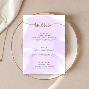 May include: A purple and gold watercolor menu with the word "menu" written in gold script. The menu lists the entree, main course, and dessert options. The entree is "Antipasto Platters", the main course is "Twice cooked shoulder of lamb, roasted vegetables" and "Honey chicken, crispy speck, green beans, buttermilk dressing", and the dessert is "Wedding Cake, Tahitian Vanilla sponge with homemade lemon curd".