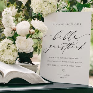 May include: White sign with black text that reads "Please sign our bible guestbook. Highlight your favorite verse and write us a note in the margin. This will become our family bible. Elyse + Adam 06.06.YY". A white bible is open on a table with white flowers.