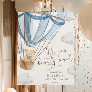 以下が含まれることがあります： 中にテディベアがいる青と白の熱気球。気球は雲のある空を飛んでいます。「We can bearly wait」というテキストが茶色の筆記体で書かれています。テキストの下には「Welcome to Hayley's Baby Shower 06.06.YY」とあります。