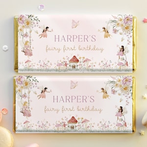 May include: Two pink and white chocolate bar wrappers with gold foil accents. The wrappers feature a fairy theme with illustrations of fairies, flowers, mushrooms, and butterflies. The text on the wrappers reads "HARPER'S fairy first birthday".