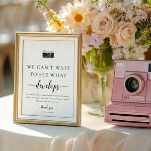 May include: A framed sign with gold trim and the text "WE CAN'T WAIT TO SEE WHAT develops" sits next to a pink instant camera. The sign includes instructions for use and a thank you message. A bouquet of flowers is in the background.