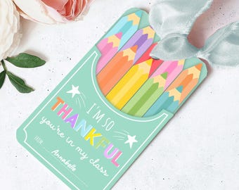 Może przedstawiać: Miętowa zielona etykieta na prezent z kolorowym wzorem ołówków. Etykieta głosi "I'm so thankful you're in my class" z imieniem "Anabelle" poniżej. Do etykiety przymocowana jest jasnoniebieska wstążka.