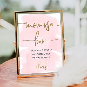 May include: A pink and gold sign with the text "mimosa bar" and instructions for making a mimosa: "Pour some bubbly, add some juice, top with fruit, cheers!"